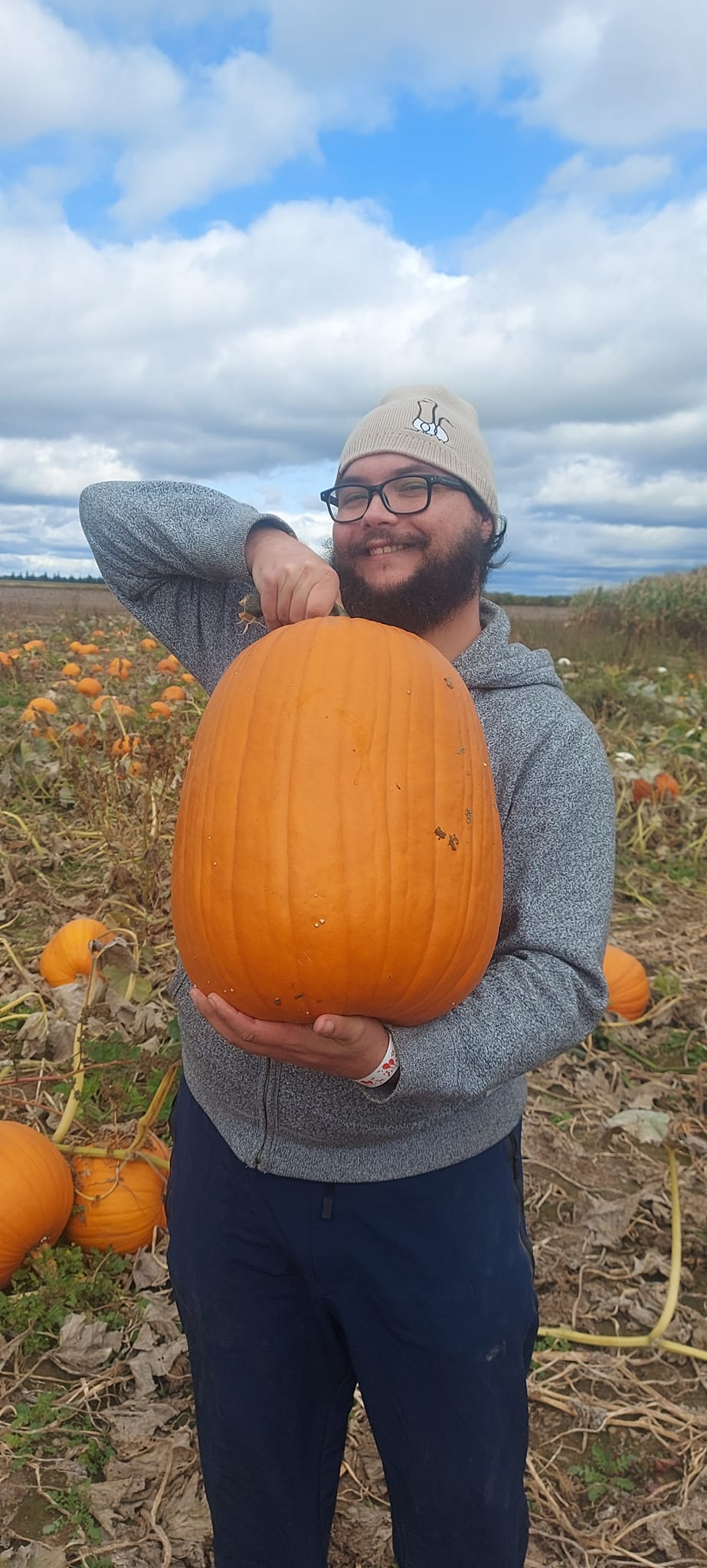 A man with short hair and bushy facial hair struggles to hold up a large, tall pumpkin roughly the size of his torso